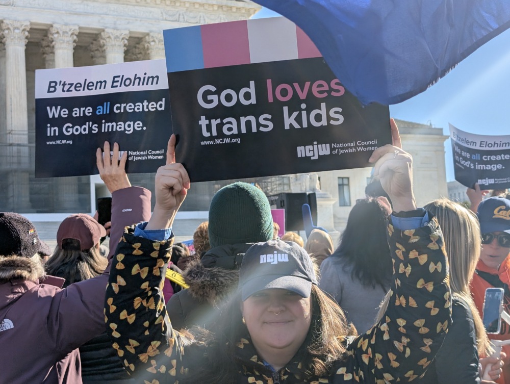 Person in an NCJW hat holds a sign in front of a rally at the Supreme Court. The sign reads "God loves trans kids."
