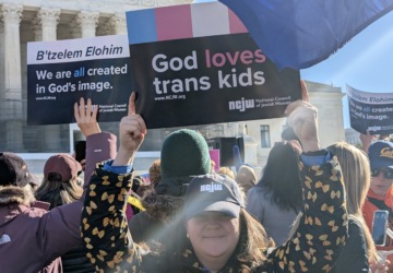 Person in an NCJW hat holds a sign in front of a rally at the Supreme Court. The sign reads "God loves trans kids."